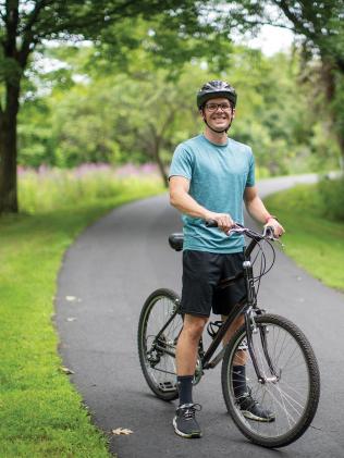 Fit man in his 30s stands astride a mountain bike on a scenic campus trail flanked by grass, flowers, trees and woods in the background