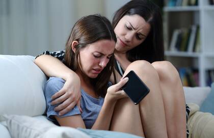 Empathetic adult comforts crying teen who holds her phone despondently.