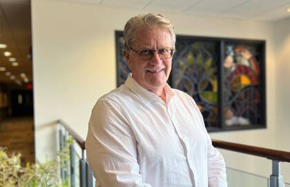 John Willard, a white man with grey hair and glasses, poses for a photo wearing a white button-down shirt.