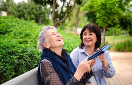Two women sitting on a bench, holding a tablet and laughing