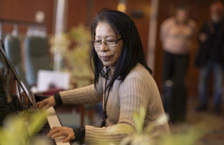 Volunteer Sylvia Ryder plays the piano in Cheshire Medical Center's lobby while people stand and listen in the blurred background