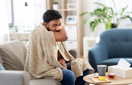 A man, sitting on a couch, covered with a blanket and coughing.