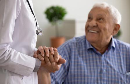 Female doctor's arm in white coat holds hand of smiling elderly man.