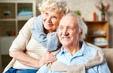 Senior man with sweater around his shoulders smiles into the distance as his wife hugs his shoulders and smiles with him.