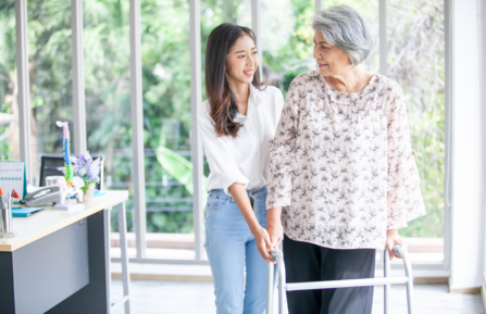 A middle aged woman uses a walker in the kitchen as her teenaged daugter assists her with care.