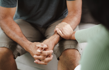 Man sits with his hands clasped pensively as a female hand reaches out to grasp his arm in comfort.