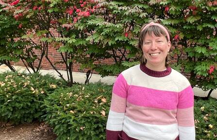 Rachael Richardson stands smiling, wearing a pink sweater and headband, in front of a decorative hedge