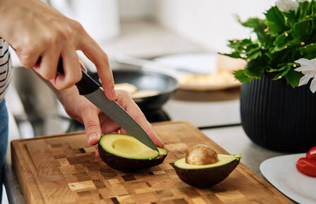 A pair of woman's hands cuts a ripe avocado that is sitting on a wooden cutting board