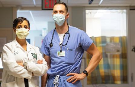 Young woman in white lab coat with surgical mask and folded arms stands beside a tall white man in scrubs with a stethoscope with hands on hips, in hallways in front of an ICU room