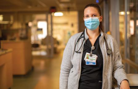 Female nurse with hair tied back, wearing surgical mask, pauses to look at camera in a dimly-lit ICU hallway