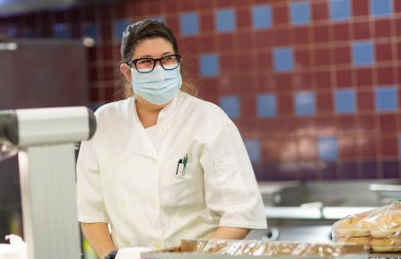 young brunette wearing a chef's coat smiles through her mask and glasses behind the counter in the cafe