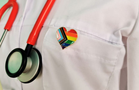 A doctor's white coat and stethescope with a pride flag heart pin on lapel