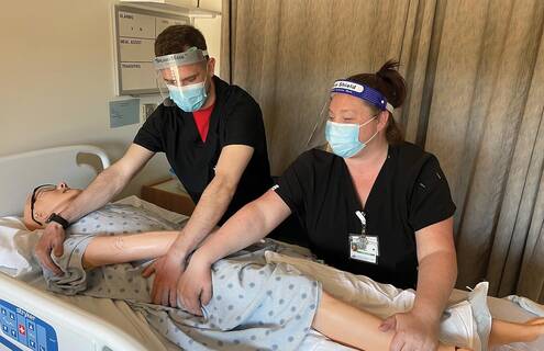 Two nurse assistant trainers wearing masks turn a mannequin on a bed