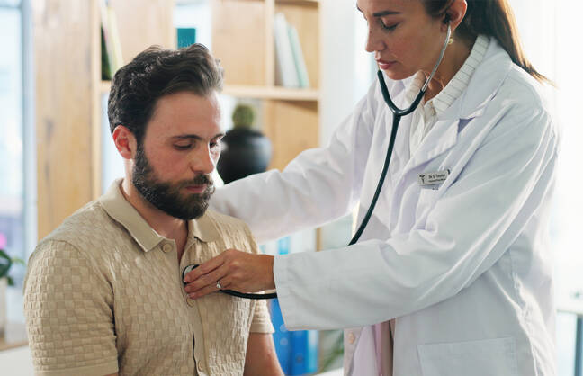 A doctor checks a patient's heart with a stethescope in an exam room.